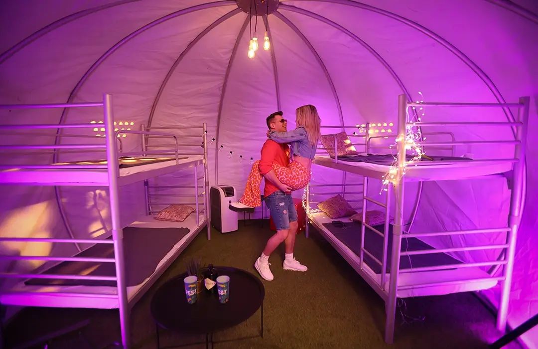 Innenraum einer Igloo Deluxe Festivalunterkunft in violettem Licht: zwei Etagenbetten, mobile Klimaanlage, kleiner Tisch und ein Paar
