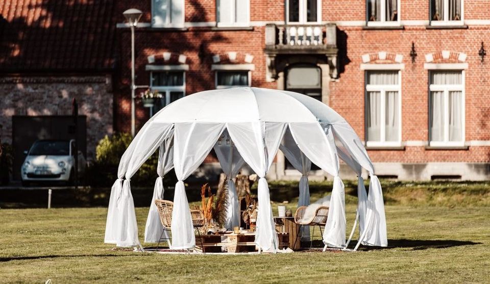 Árnyékoló borítású fehér igloo boho berendezéssel egy téglaház előtti füves kertben