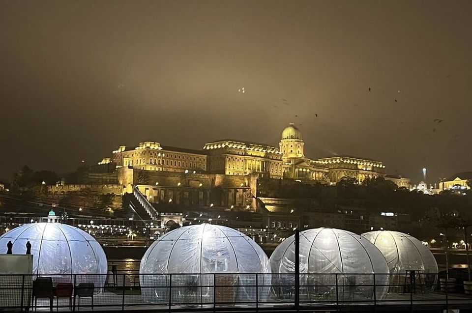 Négy igloo a pesti Duna-parton éjszaka, szemben a kivilágított budai Vár