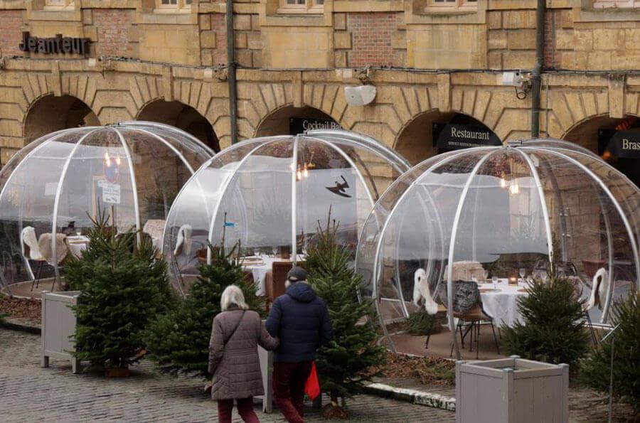 Három polikarbonát igloo terített asztalokkal egy étterem árkádjai előtt télen, fenyőkkel