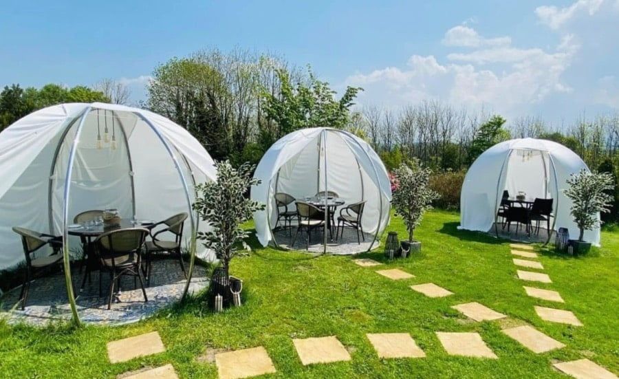 Három fehér borítású igloo kerti asztalokkal egy füves kertben, kőlapos ösvénnyel